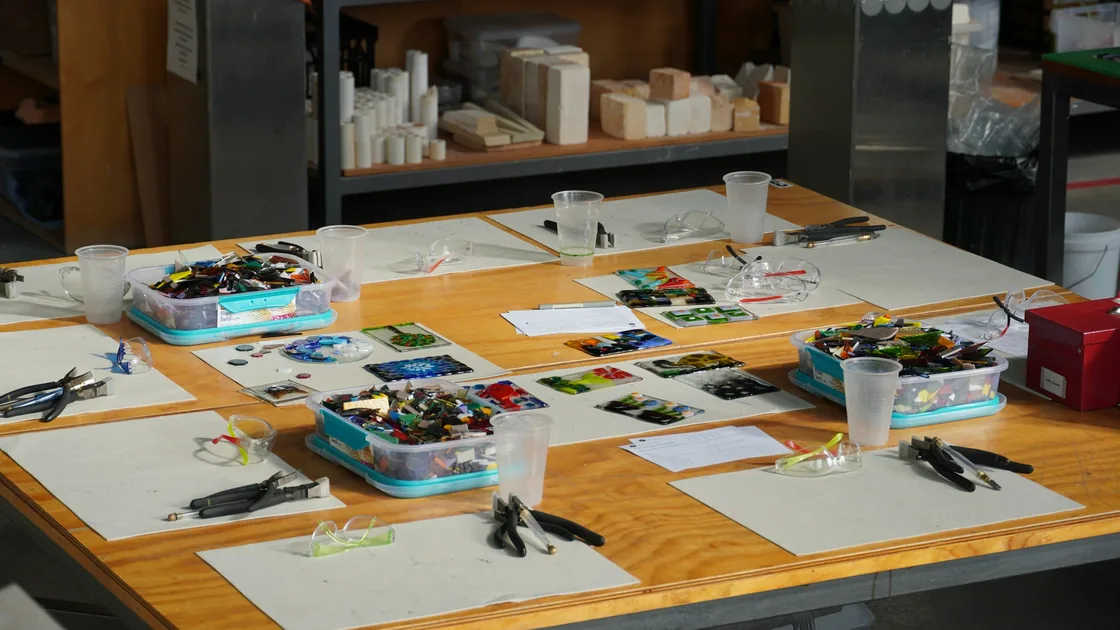 Mesa de taller con recortes de vidrio de colores en bandejas, tenazas de corte, placas refractarias y piezas planas listas para la cocción